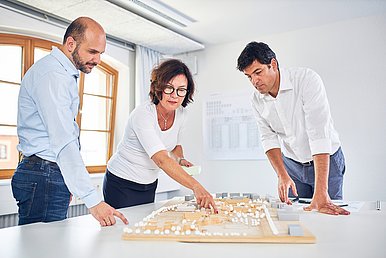 Wiebke Küpper steht mit zwei männlichen Kollegen um einen weißen Tisch, auf dem ein architektonisches Modell liegt. An der weißen Wand im Hintergrund hängt ein Bauplan.