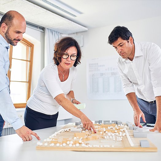 Wiebke Küpper steht mit zwei männlichen Kollegen um einen weißen Tisch, auf dem ein architektonisches Modell liegt. An der weißen Wand im Hintergrund hängt ein Bauplan.