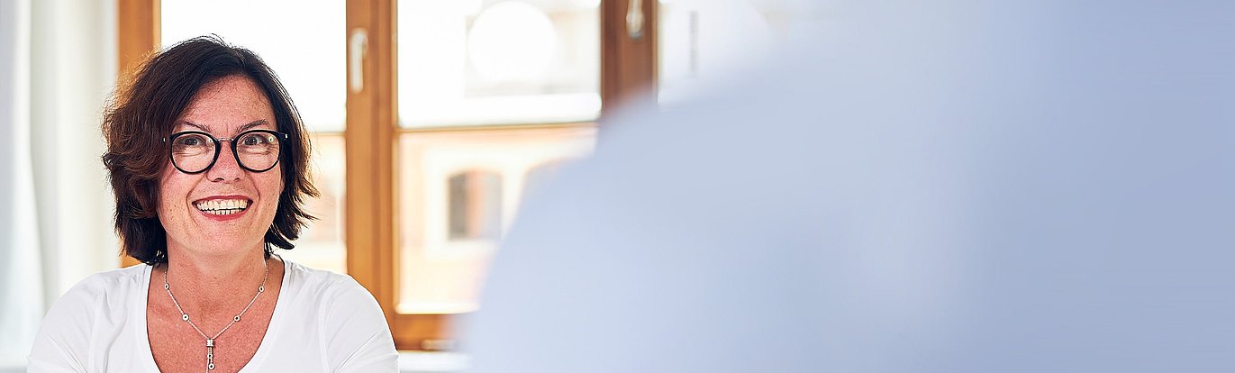 Portrait von Wiebke Küpper in einem hellen Raum vor einem Fenster. Im Vordergrund befindet sich die Schulter ihres Gesprächpartners.