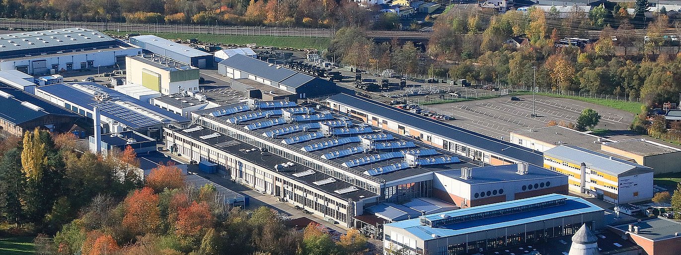 Luftbild des St. Wendeler Werks der Heeresinstandsetzungslogistik. In den Dächern einiger Gebäude spiegelt sich die Sonne. Die Anlage ist umgeben von bunt belaubten Bäumen, dem Flusslauf der Blies und einer Gleisstrecke der Deutschen Bahn.