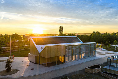 Holzgebäude des Klimapavillons mit Photovoltaikanlage auf dem Dach bei tief stehender Sonne.