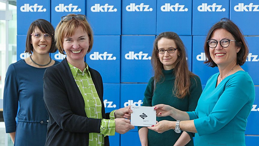 Vier strahlende Frauen stehen vor einer blauen Wand und halten eine Auszeichnung in die Kamera.