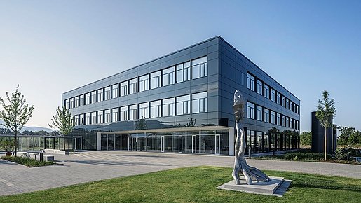 Außenaufnahme des Julius-Kühn-Instituts in Dossenheim mit klarer Architektur und langen Fensterfronten vor blauem Himmel. Vor dem Neubau steht eine graue Skulptur auf grünem Rasen. Rundherum wachsen Bäume. 