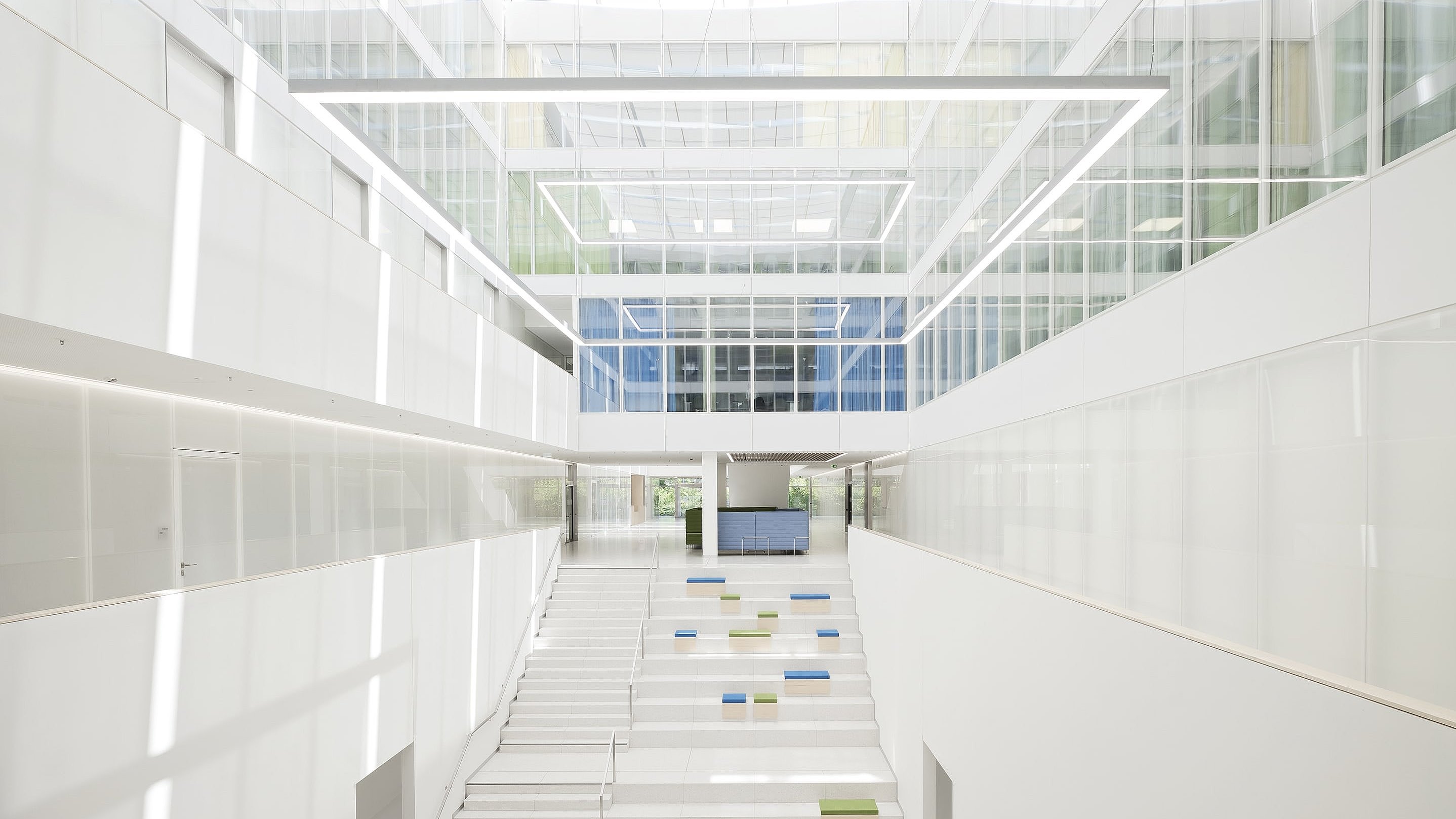Blick ins komplett in Weiß gehaltene, geschossübergreifende Atrium des Radiologischen Entwicklungszentrums in Heidelberg. Auf den Treppenstufen liegen einzelne blaue und grüne Sitzpolster.